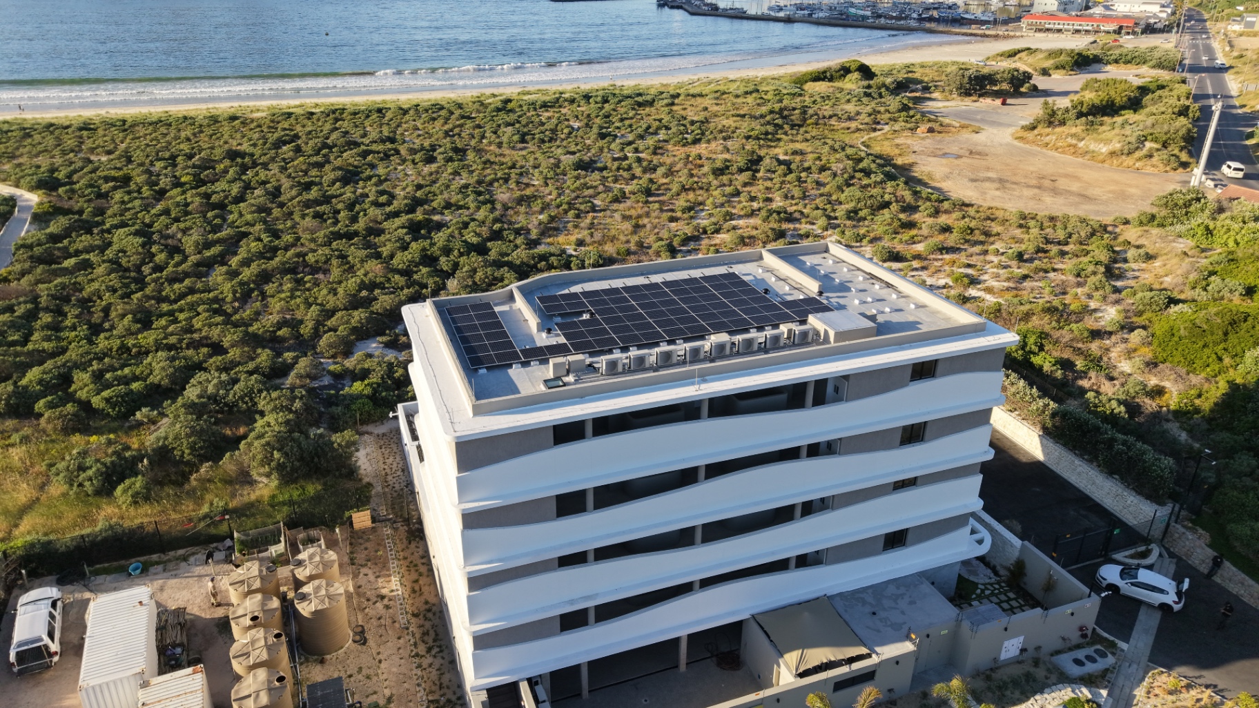 Drone view of a multi-storey beachfront building in Hout Bay with a completed solar array on its flat roof, the bay and harbour behind