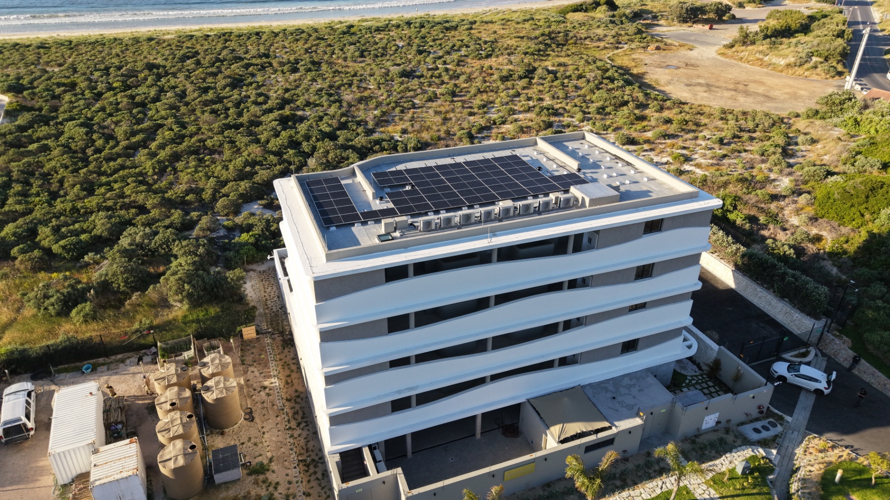 Angled drone view of a completed solar array on a flat roof with rainwater tanks and the beach beyond