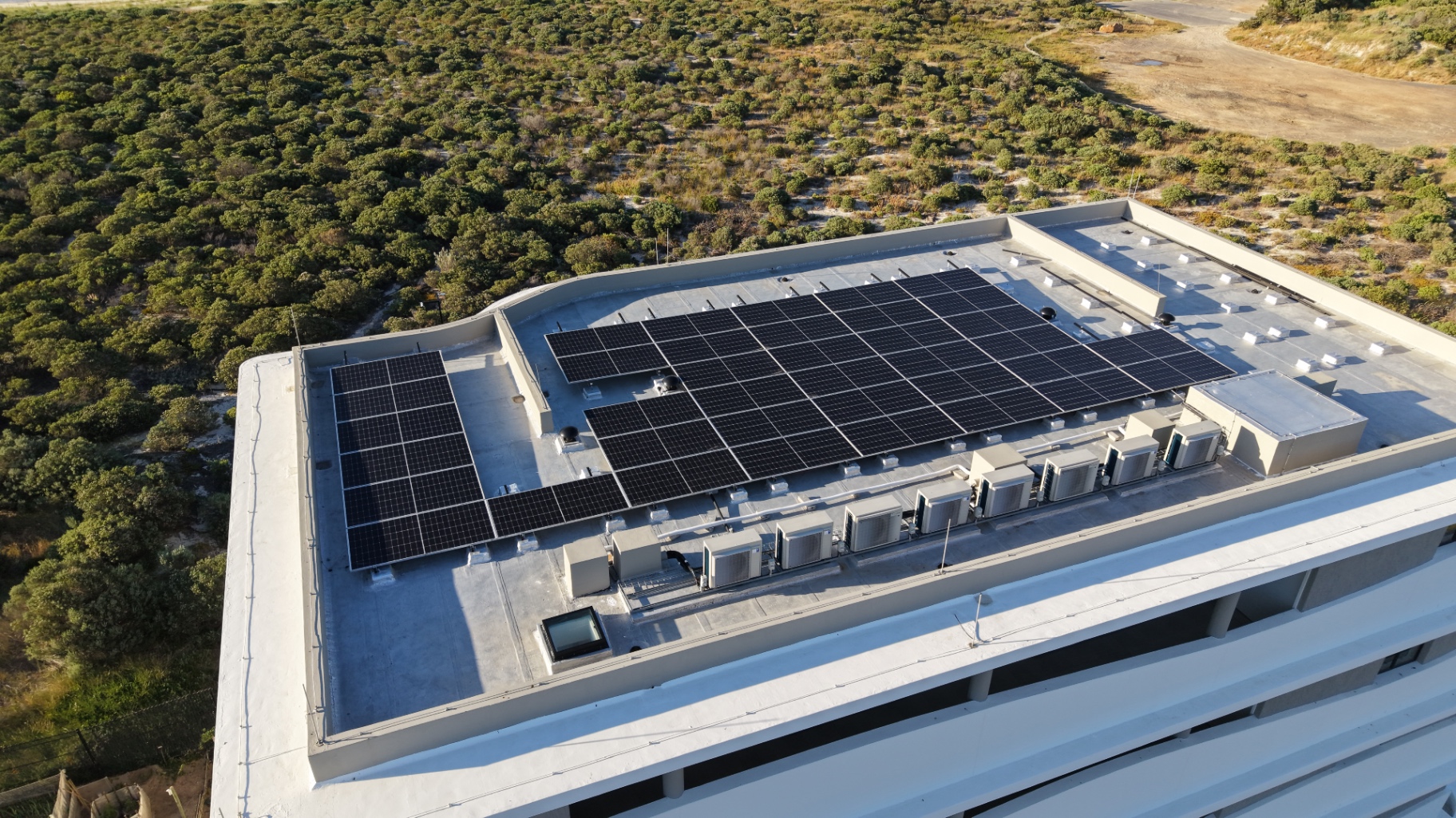 Top-down drone view of solar panels in tilted rows on a flat roof beside a line of air-conditioning condenser units
